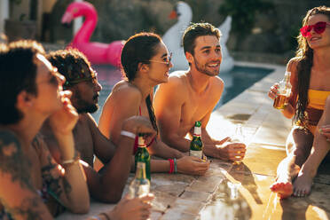 Group of men and women talking and having drinks at pool party. Friends enjoying a summer day at poolside. - JLPSF25659