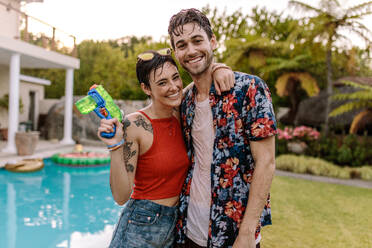 Fully wet couple standing by the swimming pool with a water gun. Beautiful women with her boyfriend standing by the pool. - JLPSF25663