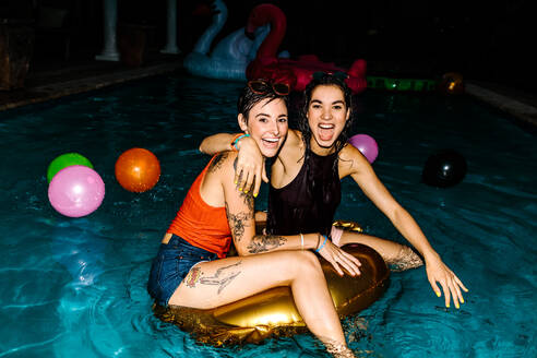 Zwei Frauen im Schwimmbad mit aufblasbarem Ring am Abend. Freundinnen genießen abendliche Poolparty. - JLPSF25664
