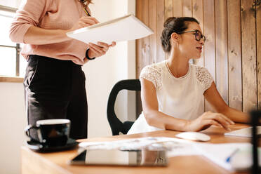 Eine Geschäftsfrau arbeitet an ihrem Schreibtisch, während eine Kollegin mit einem Notizblock daneben steht. Zwei weibliche Büroangestellte bei der Arbeit. - JLPSF25669