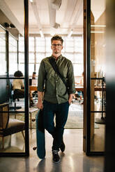 Full length shot of young man with skateboard standing in doorway of startup office with people working in background. - JLPSF25677