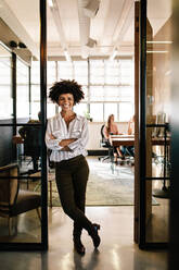Ganzkörperporträt einer erfolgreichen jungen Frau, die mit verschränkten Armen an der Bürotür lehnt. Lächelnde weibliche Führungskraft in einem Startup-Unternehmen mit arbeitenden Menschen im Hintergrund. - JLPSF25679