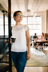 Portrait of beautiful young woman standing in office doorway with coffee. Female executive having coffee break with colleagues working in background. - JLPSF25683