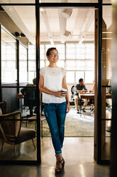 Full length portrait of smiling caucasian woman standing in office doorway with coffee and looking away. Female having coffee break at startup. - JLPSF25684