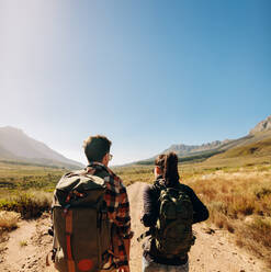 Rear view shot of young man and woman walking on a dirt road. Young couple with backpack hiking on an extreme terrain. - JLPSF25691