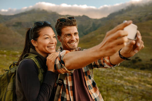Ein lächelnder junger Mann und eine Frau machen ein Selfie auf einer Wanderung. Ein junges Paar wandert im Naturschutzgebiet und macht Fotos mit dem Handy. - JLPSF25716