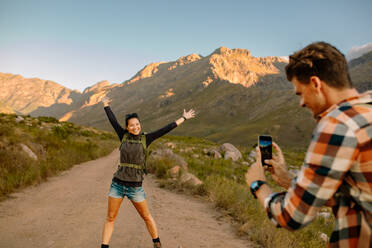 Young man taking photo of young beautiful woman standing on hiking trail with her hands raised in excitement. Couple talking pictures on summer hiking trip n nature. - JLPSF25718