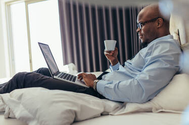 Ein Mann in formeller Kleidung arbeitet an einem Laptop, während er im Bett liegt. Ein Geschäftsmann arbeitet an einem Laptop, während er in seinem Hotelzimmer Kaffee trinkt. - JLPSF25738