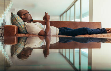 Man relaxing on a lounge and operating mobile phone. Symmetrically opposite reflection of a man relaxing on lounge falling on glass top. - JLPSF25752