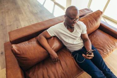 Man on a video call over mobile phone. Man wearing earphones looking at mobile phone sitting on a sofa. - JLPSF25755