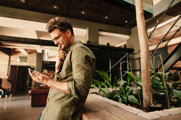 Seitenansicht eines jungen Mannes, der eine Textnachricht auf seinem Telefon liest und nachdenkt. Junger Geschäftsmann, der ein Mobiltelefon in einer Bürohalle benutzt. - JLPSF25767