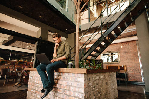 Thoughtful young man sitting in office cafeteria and looking at laptop. Executive using laptop during coffee break. - JLPSF25768