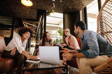 Business team sitting in office lobby and having a brainstorming session. Happy corporate business people sitting together with laptop and discussing. - JLPSF25787