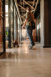 Full length rear view shot of young man skateboarding in modern office. Casual businessman skating through his startup office. - JLPSF25794