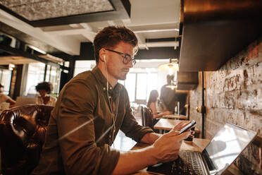 Hübscher junger Mann sitzt in einem Café mit Laptop und verwendet ein Smartphone. Lässiger Geschäftsmann liest eine Textnachricht auf seinem Handy. - JLPSF25801