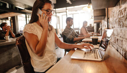 Aufnahme einer jungen Geschäftsfrau, die an einem Laptop arbeitet und mit einem Mobiltelefon in einem Startup-Büro telefoniert. Frau spricht mit einem Kunden. - JLPSF25802