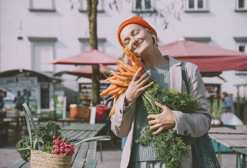 Lächelnde Frau umarmt ein Bündel Karotten auf dem Markt - NDEF00039