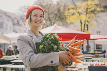 Smiling woman wearing knit hat holding fresh vegetables at market - NDEF00041