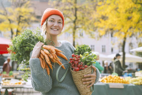 Glückliche Frau mit Gemüsesack und Karottenbündel auf der Schulter auf dem Markt - NDEF00047