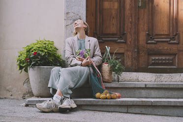 Happy woman holding cauliflower sitting with vegetables and fruit bags on staircase - NDEF00059