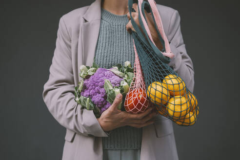 Hände einer Frau halten lila Blumenkohl und Netztaschen mit Früchten vor einer grauen Wand - NDEF00066