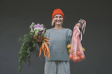 Smiling woman showing groceries in front of gray wall - NDEF00069