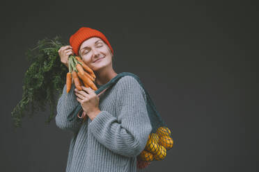 Smiling woman with eyes closed holding carrots and oranges in front of gray wall - NDEF00071