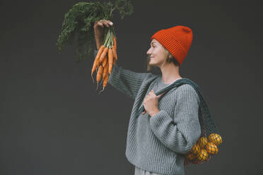 Woman carrying mesh bag looking at carrots in front of gray wall - NDEF00072