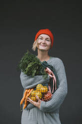 Smiling woman holding vegetables and fruits in front of gray wall - NDEF00075