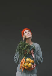 Thoughtful woman holding vegetables and fruits in front of gray wall - NDEF00076