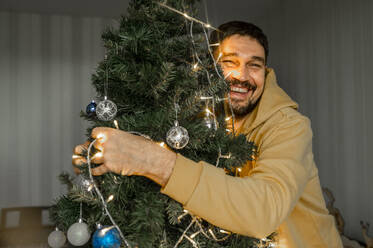 Happy man hugging illuminated Christmas tree at home - ANAF00350