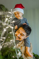 Vater trägt Sohn und schmückt Weihnachtsbaum zu Hause - ANAF00352