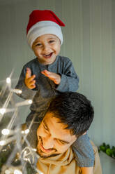 Cute boy sitting on father's shoulder decorating Christmas tree at home - ANAF00353