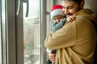 Vater umarmt Sohn in der Nähe des Fensters zu Hause - ANAF00358