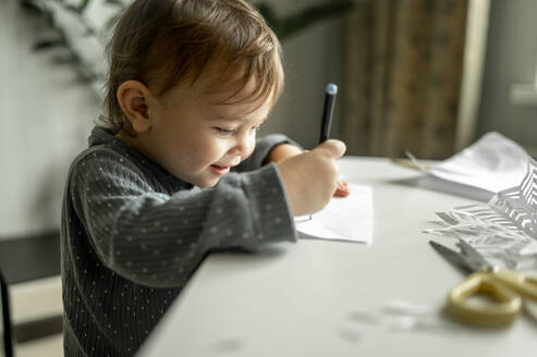 Junge zeichnet auf Papier mit Bleistift am Tisch im Haus - ANAF00371