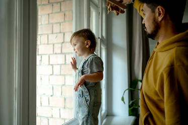 Niedlicher Junge, der durch das Fenster schaut, mit Vater, der ein Flugzeugspielzeug zu Hause hält - ANAF00398