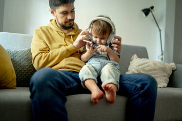 Junge benutzt sein Smartphone und hört Musik über kabellose Kopfhörer auf dem Sofa zu Hause - ANAF00399