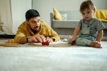 Vater und Sohn spielen mit Holzeisenbahnspielzeug auf dem Teppich zu Hause - ANAF00402