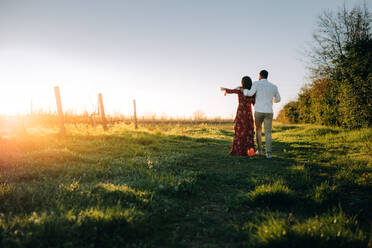 Rückansicht eines jungen Paares, das zusammen in einem Weinberg spazieren geht; die Frau zeigt dem Mann etwas im Freien. - JLPSF25818