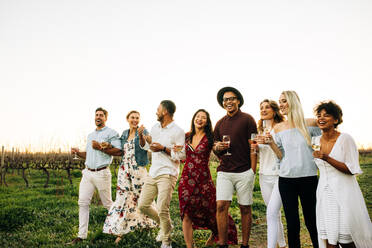 Eine Gruppe von Freunden genießt einen Spaziergang in der Natur. Multi-ethnische Millennials mit Getränken feiern im Freien in einem Weinberg. - JLPSF25821