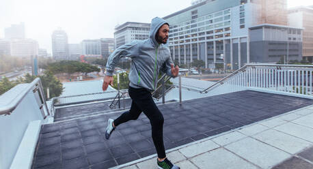 Mann, der beim Training die Treppe eines Gebäudes hinaufläuft. Männlicher Sportler in Kapuzenpulli beim Laufen in der Stadt. - JLPSF25830