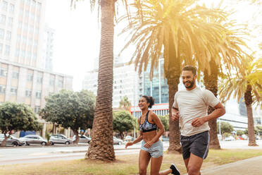 Young man and woman in jogging outfit running together outdoors. Smiling couple running on city street with trees and buildings in the background. - JLPSF25833