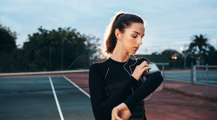 Young woman listening to music with earphones from her smart phone. Caucasian female in sportswear and sports armband at the tennis court. - JLPSF25834