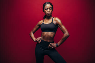 Junge Frau in schwarzer Sportkleidung vor rotem Hintergrund. Gesunde afrikanische Frau mit perfektem muskulösem Körper steht im Studio. - JLPSF25856