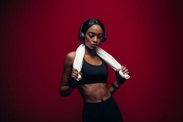 Healthy african female athlete relaxing after exercising on red background. Woman in sportswear with towel around her neck and listening to music on headphones. - JLPSF25873