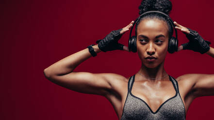 Muscular african female with headphones on red background. Fitness woman relaxing and listening music during her workout. - JLPSF25886