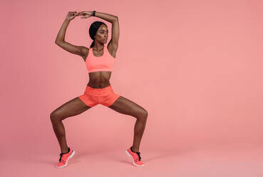 Eine fitte Frau macht Dehnungsübungen, eine Afrikanerin in Sportkleidung steht auf den Zehenspitzen vor einem rosafarbenen Hintergrund mit Copy Space. - JLPSF25925