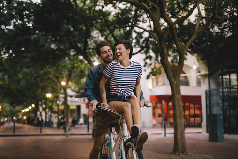 Glückliches junges Paar, das zusammen auf einem Fahrrad fährt. Frau sitzt auf dem Fahrradlenker ihres Freundes und lacht. - JLPSF25939