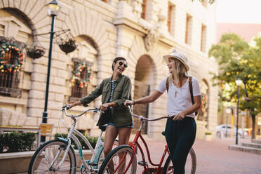 Glückliche junge Frau, die mit ihren Fahrrädern auf einer Straße spazieren geht. Weibliche Freunde, die mit Fahrrädern auf der Stadtstraße spazieren gehen. - JLPSF25941