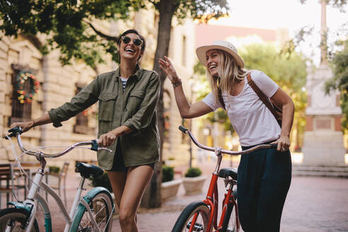 Positive und glückliche Mädchen, die mit Fahrrädern auf der Straße spazieren gehen. Weibliche Freunde, die einen Spaziergang auf der Straße mit ihren Fahrrädern genießen. - JLPSF25942